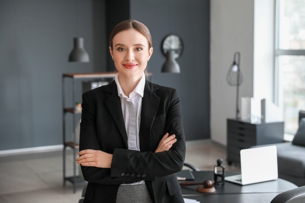 Professional woman in a dark blazer with arms crossed, smiling at the camera in a modern office, with a desk, laptop, and decorative items in the background.