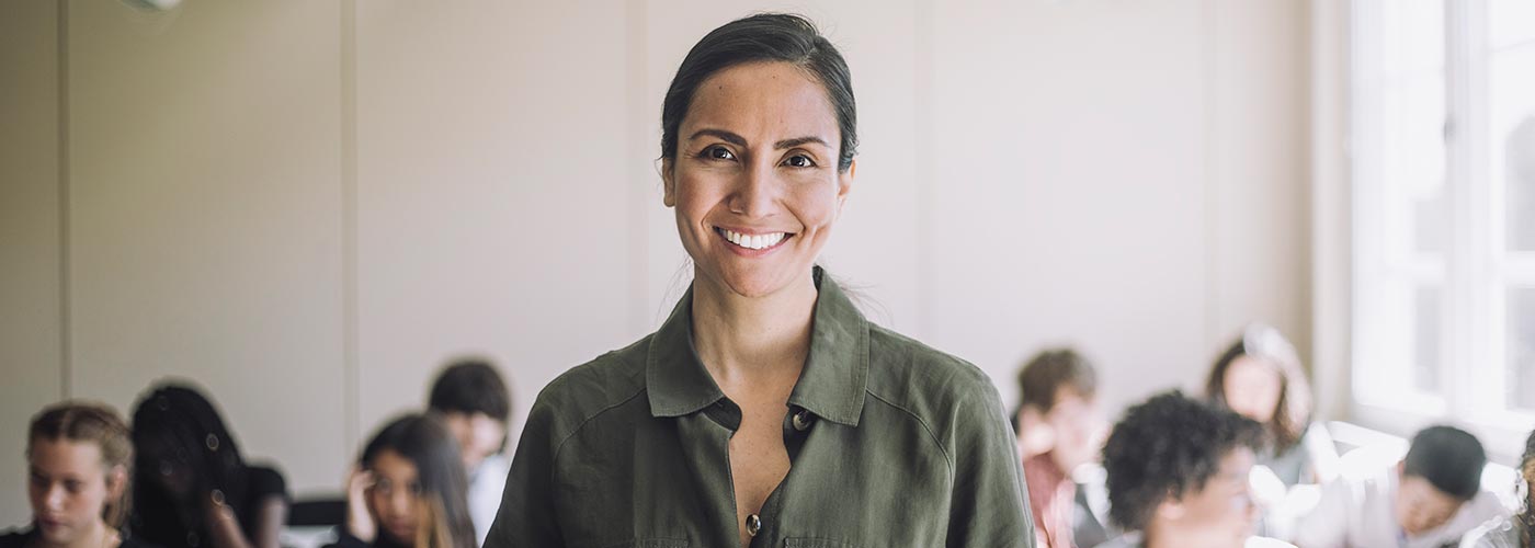 A smiling woman stands in front of a classroom, engaging with students and creating a positive learning environment.