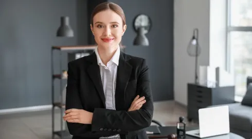 Professional woman in a dark blazer with arms crossed, smiling at the camera in a modern office, with a desk, laptop, and decorative items in the background.