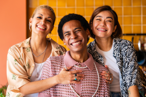Three people smiling and embracing, with a yellow tiled wall in the background.