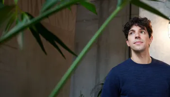 A man stands in front of a green plant, smiling and wearing a casual shirt, with a bright background.