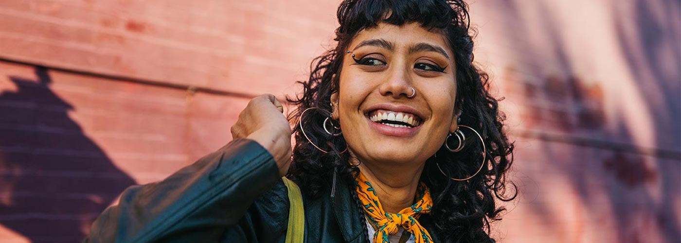 A woman with curly hair smiles brightly in front of a textured brick wall.