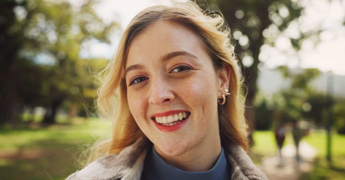 A woman smiles while standing in a lush green park, surrounded by trees and sunlight filtering through the leaves.