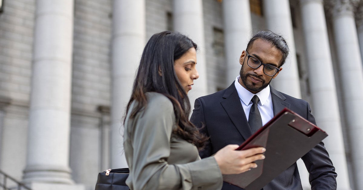 A man and woman in business attire standing outside a modern building, both smiling and engaged in conversation.